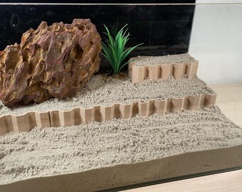 May include: A glass terrarium with a brown rock, a green plastic plant, and a brown sand substrate. The terrarium has two levels created by brown plastic edging.