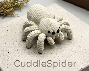 May include: A white, textured spider figurine with black eyes and eight legs. The spider is sitting on a light gray surface with the engraved text "CuddleSpider". The spider's body is round and the legs are curved.