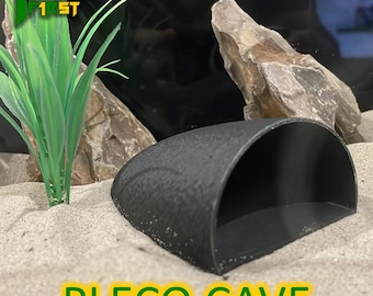 May include: Black plastic Pleco cave for an aquarium. The cave is half-round and has a textured surface. The cave is sitting on a bed of sand.