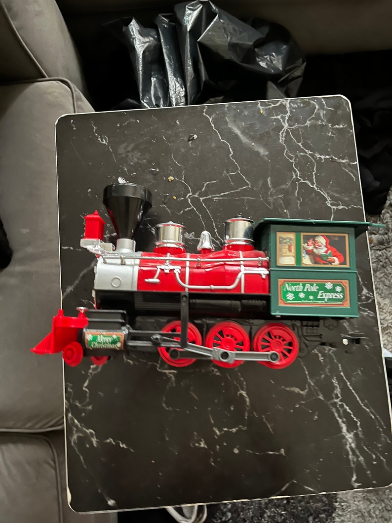 May include: A red and silver toy train engine with black wheels and a green caboose. The caboose has the words "North Pole Express" printed on it.