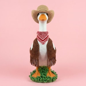 May include: A white goose figurine wearing a tan crocheted cowboy hat, a red and white patterned bandana, and a brown crocheted vest with fringe. The goose stands on a base of green crocheted grass and has orange feet and an orange beak.