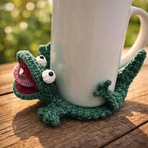 May include: A green crocheted lizard-shaped mug hugger wrapped around a plain white ceramic mug. The lizard features a wide-open mouth with a pink tongue, two white round eyes with black pupils, and textured limbs that grip the sides of the mug.