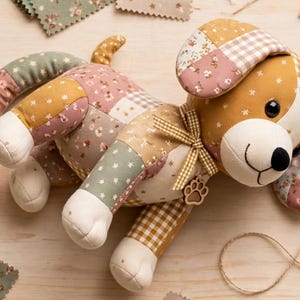 May include: A stuffed dog toy made from patchwork fabric in shades of mustard yellow, sage green, and dusty pink. The toy features floral and gingham patterns, a brown and white checkered ribbon around its neck, and a small wooden paw print charm.