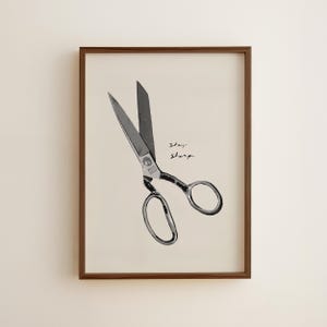 May include: A framed print featuring a pair of open scissors. The scissors are black and silver, set against a cream background. The words "Stay Sharp" are written in cursive. The frame is a dark brown color.