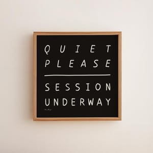 Könnte beinhalten: Ein gerahmtes schwarzes Schild mit weißem Text, der „QUIET PLEASE SESSION UNDERWAY“ lautet. Das Schild hat einen Naturholzrahmen und ist an einer weißen Wand befestigt. Der Text ist in einer einfachen, serifenlosen Schriftart.