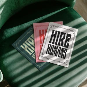 Three prints with the text HIRE HUMANS are fanned out across a dark green upholstered chair.