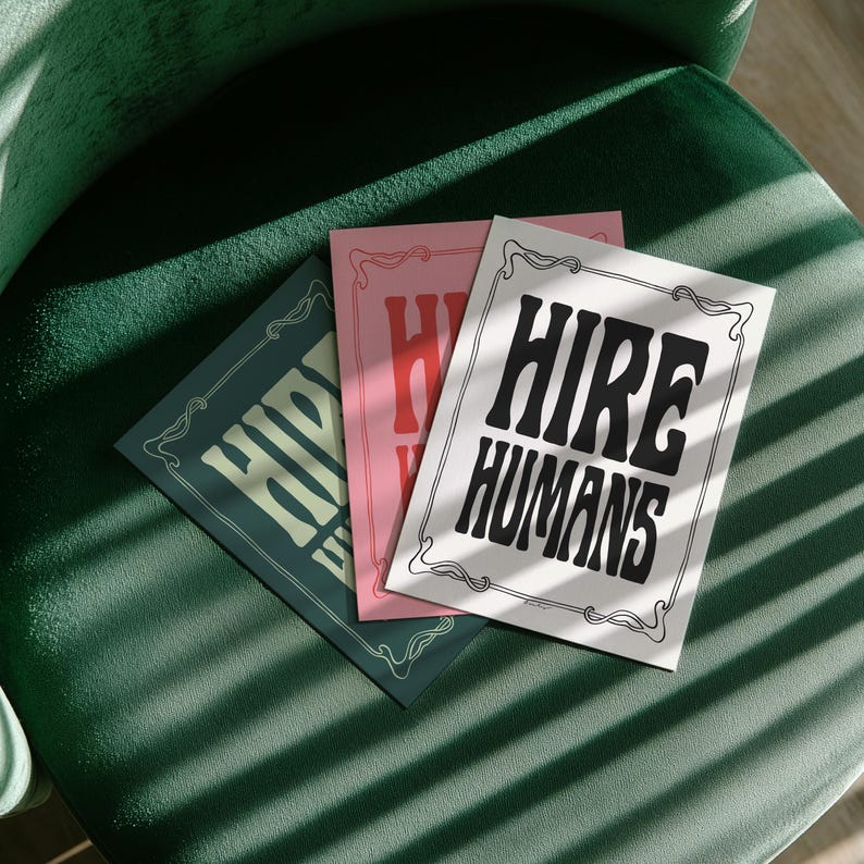 Three prints with the text HIRE HUMANS are fanned out across a dark green upholstered chair.