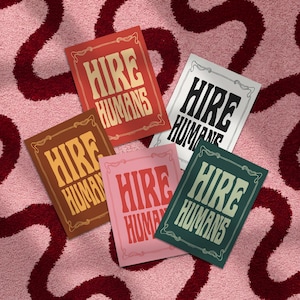 Five different colors of an art print with the words HIRE HUMANS in a hand-lettered art nouveau style are fanned out on a textured pink and burgundy patterned rug.