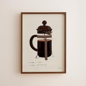 May include: A framed print featuring a brown French press coffee maker filled with dark liquid. The print has the words "works well under pressure" in a handwritten font. The frame is a dark brown color.