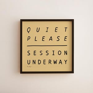 Könnte beinhalten: Ein gerahmter Kunstdruck mit dem Text "QUIET PLEASE SESSION UNDERWAY" in schwarzer, handschriftlicher Schrift auf beigem Hintergrund. Der Druck befindet sich in einem dunkelbraunen Rahmen und ist an einer weißen Wand befestigt.