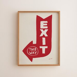 May include: A framed wall art print featuring a bold red arrow shape on a cream background. The word EXIT is printed in white capital letters vertically along the arrow, with the text THIS WAY inside a white circle at the arrow's point.