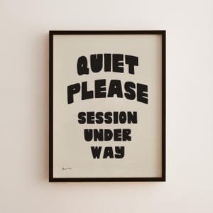 Könnte beinhalten: Ein gerahmter Druck mit den Worten "QUIET PLEASE SESSION UNDER WAY" in fetten, schwarzen Buchstaben. Der Text ist auf einem cremefarbenen Hintergrund zentriert und in einen schwarzen Rahmen gefasst. Der Druck hängt an einer weißen Wand.