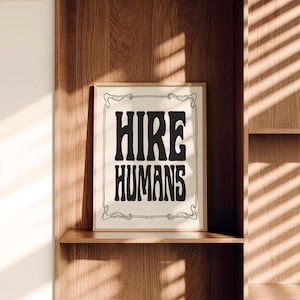 A framed black and white HIRE HUMANS art print sits upright on a wooden shelf.