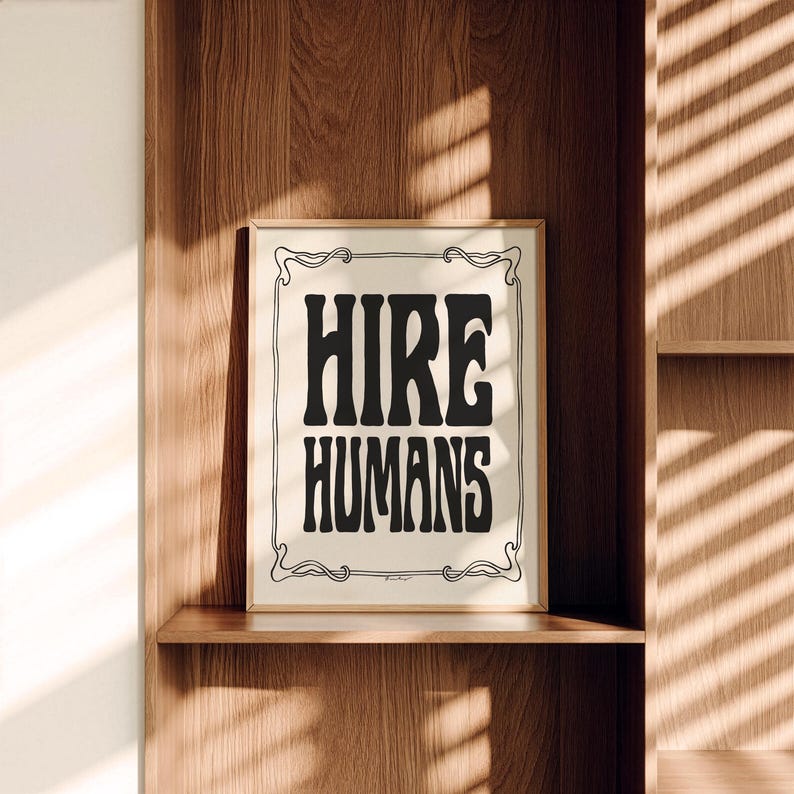 A framed black and white HIRE HUMANS art print sits upright on a wooden shelf.
