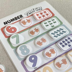 May include: A colorful educational game with the words "NUMBER MATCH" printed on the top. The game features numbered circles and matching sets of objects, including hands and various illustrations. The numbers are from 6 to 9.