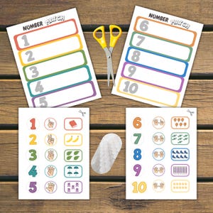 May include: Two educational worksheets titled Number Match featuring numbers one through ten, alongside two corresponding answer sheets with illustrations of hands, objects, and shapes, accompanied by yellow-handled scissors and a white oval fastener.
