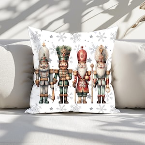May include: A white cushion with a festive design featuring four nutcracker soldiers in various poses. The background is a white snowflake pattern with silver stars.