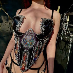 May include: A black leather corset with silver accents and stained glass-like panels featuring religious figures. The corset has a pointed bottom and is worn with a matching black garter belt.