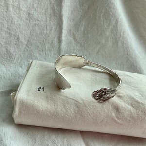 K&ouml;nnte beinhalten: Ein silbernes Armband aus einem L&ouml;ffel mit floralen Details. Das Armband ist aus einem recycelten L&ouml;ffel gefertigt und an den Enden offen. Es liegt auf einem wei&szlig;en Tuch mit der Aufschrift "#1".