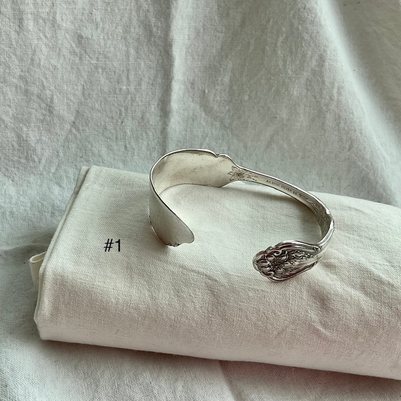 K&ouml;nnte beinhalten: Ein silbernes Armband aus einem L&ouml;ffel mit floralen Details. Das Armband ist aus einem recycelten L&ouml;ffel gefertigt und an den Enden offen. Es liegt auf einem wei&szlig;en Tuch mit der Aufschrift "#1".