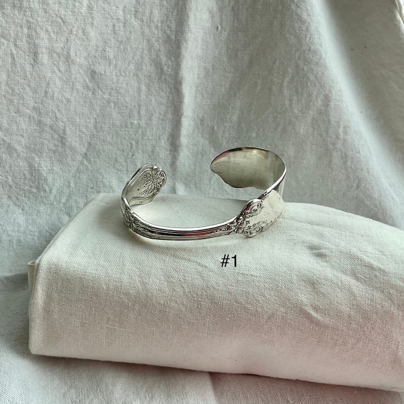 K&ouml;nnte beinhalten: Ein silbernes Armband aus einem L&ouml;ffel mit kunstvollen floralen und scrollenden Details. Das Armband ist oben offen und hat die Nummer "#1" darunter aufgedruckt. Das Armband liegt auf einem wei&szlig;en Tuch.