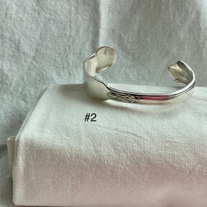K&ouml;nnte beinhalten: Ein silberfarbenes Armreif-Armband aus einer wiederverwerteten Gabel. Das Armband hat ein dekoratives Blumendesign auf der Vorderseite und die Nummer "#2" ist auf dem wei&szlig;en Stoff darunter aufgedruckt. Das Armband wird auf einem wei&szlig;en Tuch pr&auml;sentiert.