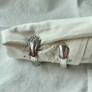 K&ouml;nnte beinhalten: Zwei silberne Serviettenringe aus recyceltem Besteck. Ein Ring hat ein verziertes Blumendesign, w&auml;hrend der andere einen stromlinienf&ouml;rmigen Art-Deco-Stil hat. Die Ringe halten eine gefaltete wei&szlig;e Stoffserviette.