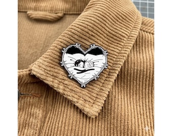 May include: A black and white heart-shaped enamel pin featuring a cartoon couple in bed, surrounded by barbed wire. The pin is set against a tan corduroy jacket, showcasing the texture of the fabric.