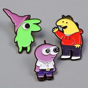 May include: Three cartoon character enamel pins. One is green with a purple hat, another is yellow in a red hoodie, and the third is purple with large eyes. All have black outlines and are on a grey background.