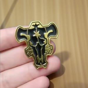 Puede incluir: Un pin metálico dorado con un emblema negro. El emblema presenta una cabeza de toro estilizada con una estrella en el centro. El pin se sostiene en una mano sobre un fondo de madera.