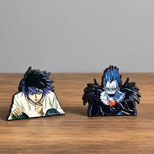 May include: Two enamel pins featuring anime characters. One pin depicts a character with black hair and a white shirt, the other shows a character with blue hair and a black outfit. Both pins are on a wooden surface.