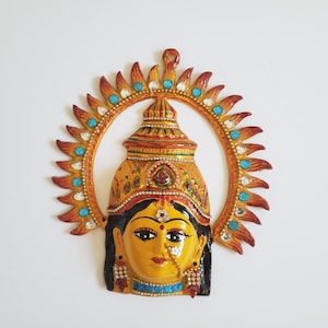 May include: A colourful, hand-painted metal wall hanging depicting a Hindu goddess with a yellow face, black hair, and a crown. The goddess is surrounded by a circular halo with red, orange, and blue accents.