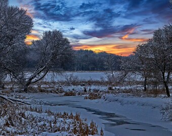 May include: A frozen river winds through a snowy forest at sunset. The sky is a vibrant mix of blue, orange, and pink. Trees are covered in snow, and the river is partially frozen.