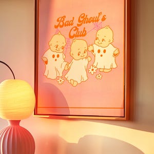 May include: Framed artwork featuring the text "Bad Ghoul's Club" in orange script above three cartoon ghost babies on a pink background. The artwork is in a wooden frame and has a retro aesthetic.