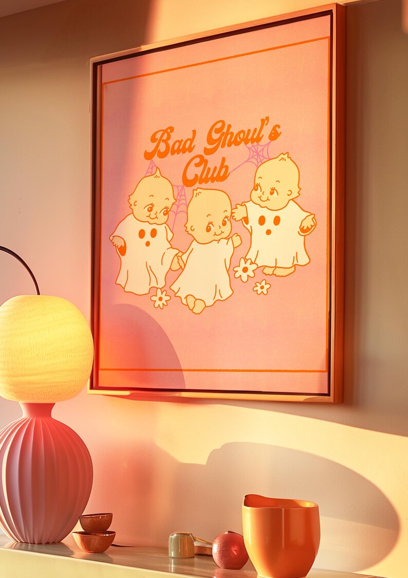 May include: Framed artwork featuring the text "Bad Ghoul's Club" in orange script above three cartoon ghost babies on a pink background. The artwork is in a wooden frame and has a retro aesthetic.