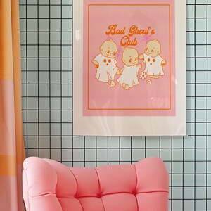 May include: A framed pink art print with the text "Bad Ghoul's Club" and illustrations of three ghost-like babies. The print is displayed on a light blue tiled wall, with a pink armchair in the foreground.
