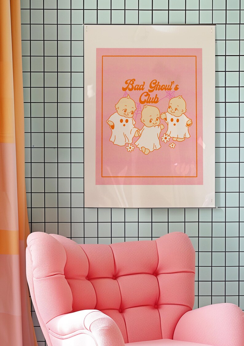 May include: A framed pink art print with the text "Bad Ghoul's Club" and illustrations of three ghost-like babies. The print is displayed on a light blue tiled wall, with a pink armchair in the foreground.