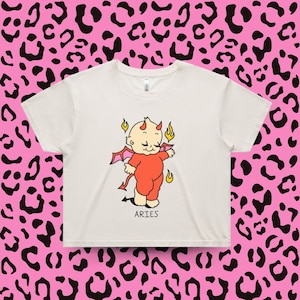 Puede incluir: Top corto color crema con un beb&eacute; diablo de dibujos animados con cuernos, alas y llamas. El beb&eacute; lleva un traje rojo. La palabra "ARIES" est&aacute; impresa debajo.