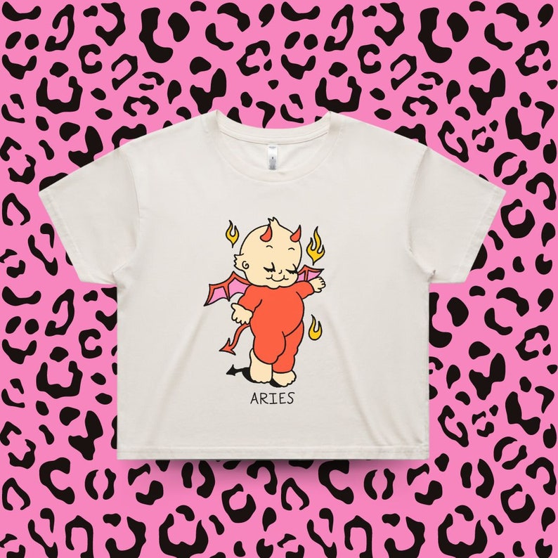 Puede incluir: Top corto color crema con un beb&eacute; diablo de dibujos animados con cuernos, alas y llamas. El beb&eacute; lleva un traje rojo. La palabra "ARIES" est&aacute; impresa debajo.