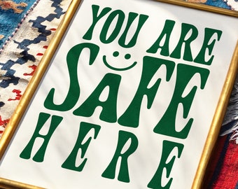 Könnte beinhalten: Ein gerahmter Druck mit einem goldenen Rahmen und weißem Hintergrund. Der Text "You are safe here" ist in grünen Buchstaben mit einem Smiley in der Mitte des Wortes "safe" geschrieben.