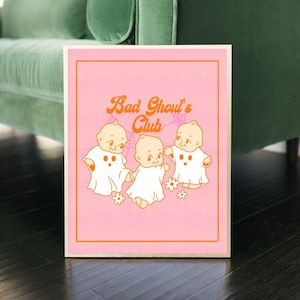 May include: A framed print featuring the text "Bad Ghoul's Club" in orange script above three cartoon ghosts. The ghosts are depicted as babies wearing white ghost costumes, set against a pink background with an orange border.
