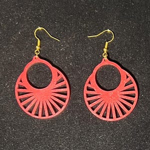 May include: A pair of red wooden earrings with a sunburst design. The earrings have a circular shape with a cut-out circle in the center and gold-colored ear wires. The earrings are displayed against a dark background.