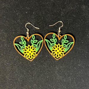 May include: A pair of heart-shaped earrings with a sunflower design. The earrings feature a yellow sunflower in the center, surrounded by green leaves and a brown border. The earrings have silver hooks.