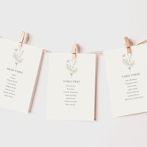 Könnte beinhalten: Drei weiße Karten mit floralen Illustrationen und Text, der die Namen für einen Hochzeitsempfang auflistet. Die Karten sind mit "Head Table", "Table Two" und "Table Three" beschriftet.