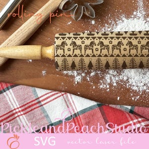 May include: A wooden rolling pin with a brown and black design of Scandinavian horses and Christmas trees. The rolling pin is on a wooden surface with white flour scattered around it. The text "rolling pin" is written in pink on the top of the image. The text "Pick and Peach Studio SVG vector laser file" is written in pink at the bottom of the image.