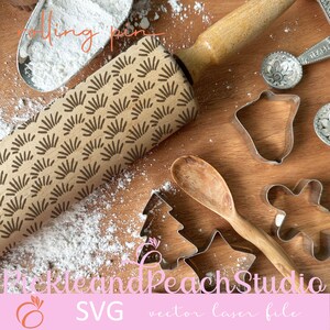 May include: A wooden rolling pin with a brown geometric pattern. The rolling pin is surrounded by cookie cutters, a wooden spoon, and powdered sugar on a wooden surface. The text "Pickle and Peach Studio SVG vector laser file" is visible at the bottom of the image.