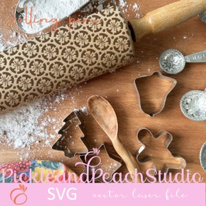 Peut inclure: Un rouleau à pâtisserie en bois avec un motif de flocon de neige, une cuillère en bois et des emporte-pièces en métal en forme de cloche, d'étoile et d'arbre. Les emporte-pièces sont sur une surface en bois avec du sucre en poudre éparpillé autour d'eux. Le texte "Pickle and Peach Studio SVG vector laser file" est en bas de l'image.