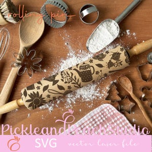 Puede incluir: Un rodillo de madera marrón con un diseño floral y una tetera. El rodillo está sobre una superficie de madera con harina esparcida a su alrededor. También hay cortadores de galletas y una pequeña pala de harina en la imagen.