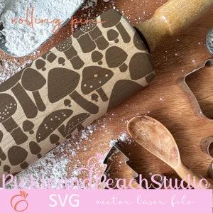 May include: A wooden rolling pin with a brown mushroom design. The rolling pin is on a wooden surface with white powder and a wooden spoon. The text "rolling pin" is written in pink on the top of the image. The text "Pickle and Peach Studio" is written in pink at the bottom of the image. The text "SVG vector laser file" is written in black at the bottom of the image.