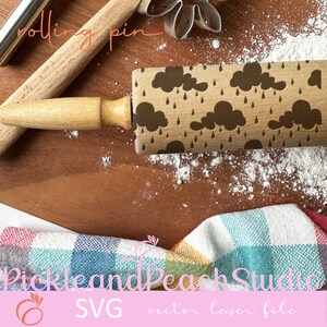 Pode incluir: Um rolo de massa de madeira com um design marrom e preto de nuvens e gotas de chuva. O rolo está sobre uma superfície de madeira com farinha espalhada ao redor. O texto "rolling pin" está escrito em preto sobre um fundo branco acima do rolo.